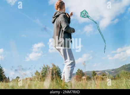 Teenager boy with flying green color kite on the high grass meadow in the mountain fields. Happy childhood moments or outdoor time spending concept im Stock Photo