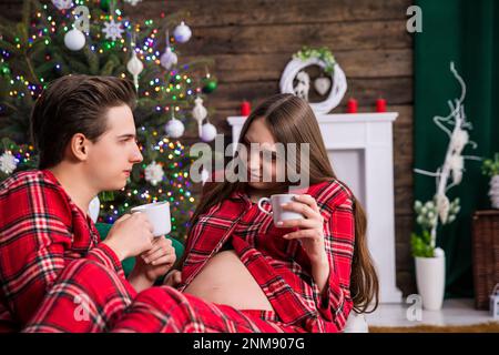 A couple expecting a baby sits on a gray couch against a backdrop of a dressed Christmas tree with lights. The man is holding the woman on his lap, bo Stock Photo