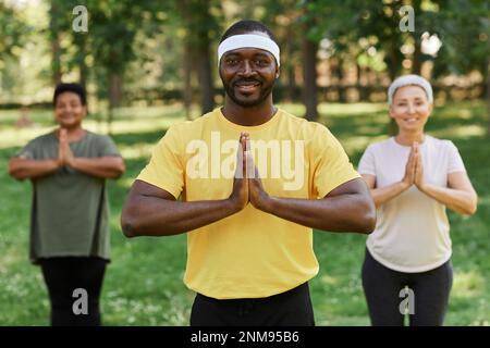 Waist up portrait of male yoga instructor smiling at camera in park with senior women in background Stock Photo