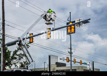Worker up high in crane bucket repairing traffic light at city intersection on cloudy day with building in background Stock Photo