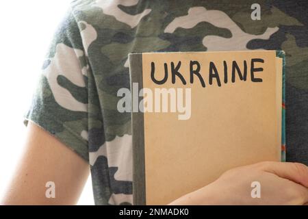 A girl in a camouflage uniform with a book on which Ukraine is written, education in Ukraine during the war, student Stock Photo