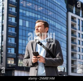 Man holding mobile phone in his hands standing on street against urban building. Stock Photo
