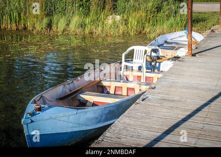 Metal row boats - one with plastic chair in it- pulled up to pier with mossy weedy pond bank behind. Stock Photo