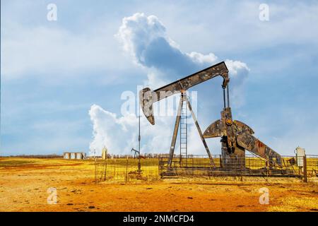 Pump jack oil gas well on red soil with storage tanks on horizon under dramatic sky Stock Photo