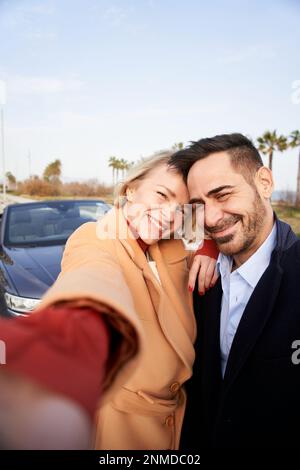 Vertical happy middle-aged couple take selfie outside car. Wife and husband look smiling at camera.  Stock Photo