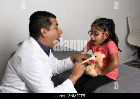 Latino pediatrician doctor checks his girl patient with autism spectrum disorder ASD, he communicates through his teddy bear Stock Photo