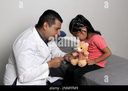 Latino pediatrician doctor checks his girl patient with autism spectrum disorder ASD, he communicates through his teddy bear Stock Photo