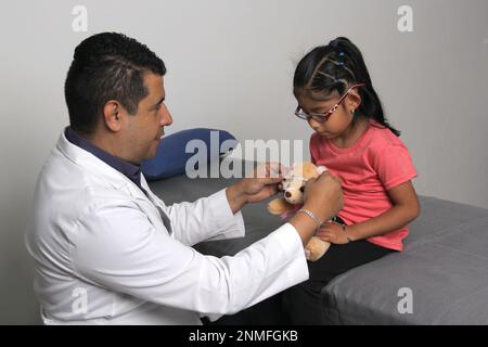 Latino pediatrician doctor checks his girl patient with autism spectrum disorder ASD, he communicates through his teddy bear Stock Photo