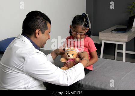 Latino pediatrician doctor checks his girl patient with autism spectrum disorder ASD, he communicates through his teddy bear Stock Photo