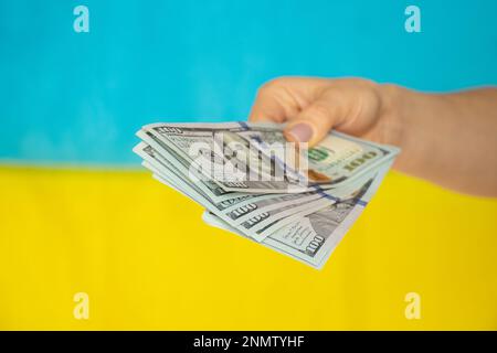 A woman's hand gives money in dollars against the background of the flag of Ukraine, financial support for Ukraine and money Stock Photo