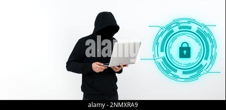 Male programmer in black hoodie stands and holds laptop. Hacker with hood on his head typing text on computer. Futuristic digital design on the wall. Stock Photo