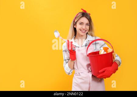 Young housewife with bucket of household items and brush on yellow background. Space for text Stock Photo