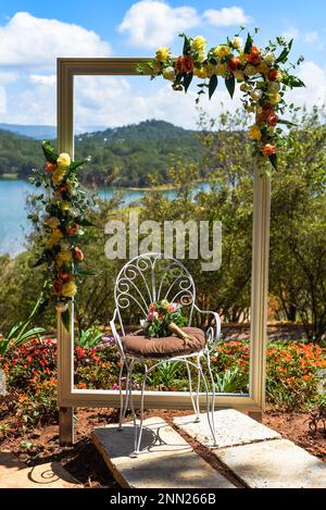 Photo zone for wedding ceremony with decorated frame and chair with flowers without people against landscape with lake Stock Photo