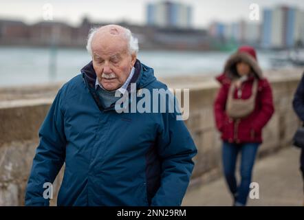 A 96-year-old man looks down as he carefully walks alongside the sea wall at Portsmouth Harbour, UK. Stock Photo