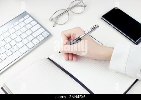 A young female hand writes with a pen in a checkered notebook with a pen, a keyboard, glasses and a smartphone lie nearby.  On the white table Stock Photo
