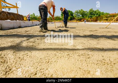 Construction workers, riggers are using rake and wooden handmade handcraft tool to spreading, leveling concrete covering square trench, pouring layer Stock Photo