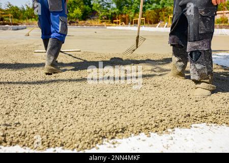 Construction workers, riggers are using rake and wooden handmade handcraft tool to spreading, leveling concrete covering square trench, pouring layer Stock Photo