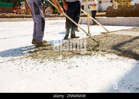 Construction workers, riggers are using rake and wooden handmade handcraft tool to spreading, leveling concrete covering square trench, pouring layer Stock Photo