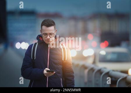 Man looking at phone while walking on busy city street. Urban scene with cars at twilight. Stock Photo