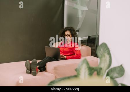 Full length photo of gorgeous curly haired business woman dressed in red shirt, black pants and red boots lying on a pink sofa at modern office workin Stock Photo
