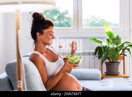 Pretty young woman eating salad on sofa at home. Healthy lifestyle during maternity period Stock Photo
