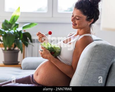 Pretty young woman eating salad on sofa at home. Healthy lifestyle during maternity period Stock Photo