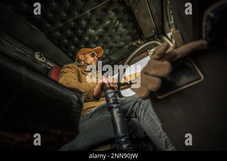 Professional Caucasian Trucker Driving His American Semi Truck. Bottom View from Inside of the Cabin. Stock Photo