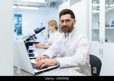 Portrait of mature successful scientist lab technician at workplace in laboratory with microscope, senior man with beard and goggles smiling and looking into camera, inventor with laptop . Stock Photo