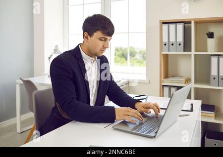 Young man working in office, sitting at his desk and typing on modern laptop computer Stock Photo