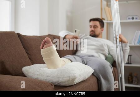 Man with an injured, broken leg relaxing on the sofa, with his foot resting on a cushion Stock Photo