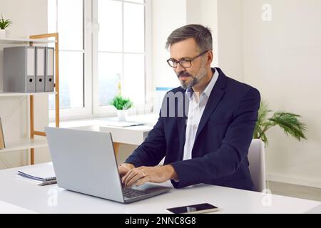 Happy mature businessman sitting at his working desk and typing on his laptop computer Stock Photo