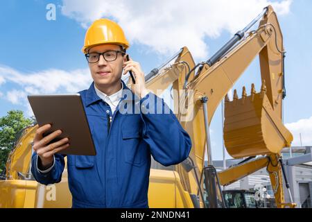 Engineer in a helmet with a digital tablet and phone on the background of construction machines Stock Photo