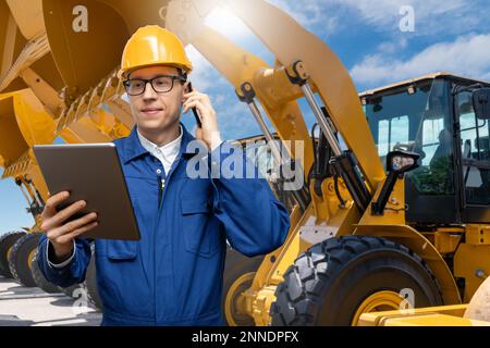 Engineer in a helmet with a digital tablet and phone on the background of construction machines Stock Photo