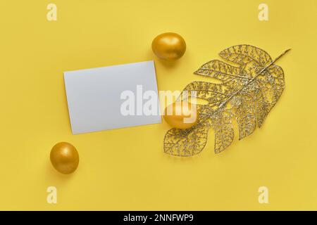 Gold eggs, monstera leaf from wire, blank paper, yellow background. Stock Photo