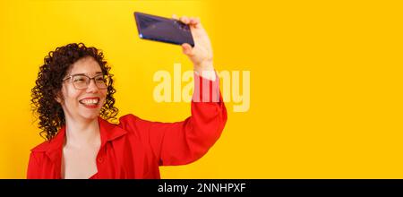 Banner with pretty curly girl with glasses taking a selfie. A young woman takes a picture of herself on the phone. Studio female portrait on a bright Stock Photo