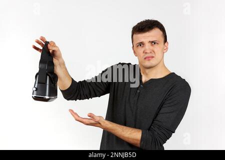 A young man holds a virtual reality helmet in his hands and looks questioningly into the frame. The figure is isolated on a white background. Stock Photo