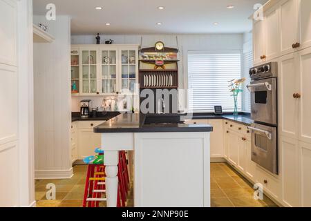 White painted shaker style cabinets and island with South African black marble countertop and red and white painted folk art barstools in kitchen. Stock Photo