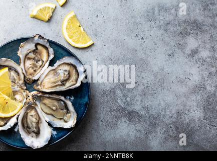 Set of half dozen fresh opened oysters in shell with lemon wedges served on rustic blue plate on gray stone background, close up, top view, space for Stock Photo