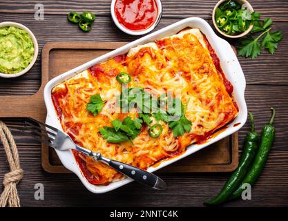 Traditional Mexican dish enchiladas with meat, chili red sauce and cheese in white casserole dish over rustic wooden background, served with Stock Photo