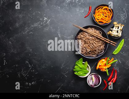 Ingredients for making stir-fried noodles soba. Cut fresh vegetables, mushrooms, boiled soba noodles in bowl with chopsticks ready for cooking, black Stock Photo