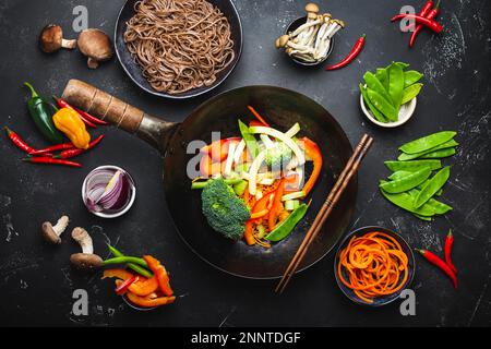 Ingredients for making stir-fried noodles soba. Cut fresh vegetables in wok pan, boiled soba noodles in bowl with chopsticks ready for cooking, black Stock Photo