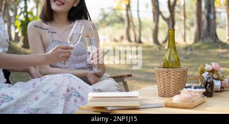 Two beautiful Asian women in lovely dresses enjoying picnic in the park, sipping white wine, sitting on picnic chairs Stock Photo