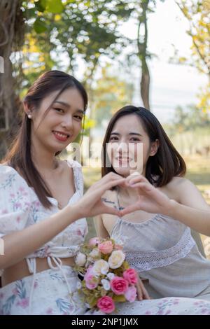 Asian woman friends, they are having picnic,they talk happily,charming Asian woman makes a heart hand sign with her friend while picnic in the park Stock Photo