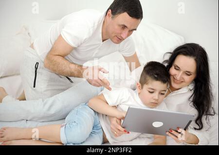 people, family and technology concept mother father and little boy with tablet pc computer in bed at home or hotel room. Happy family spend time together lying under white blanket on bed Stock Photo