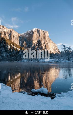 The rocky surface of the Sierra Nevada mountains, reflecting early morning sunlight, being reflected in Merced river, on an early winter, snow-covered Stock Photo