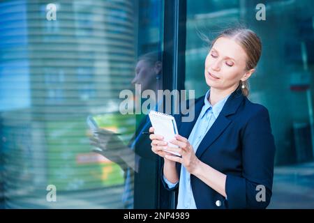Successful beautiful business woman standing against backdrop buildings writes notes in a notebook or plans a working day in business clothes. Stock Photo