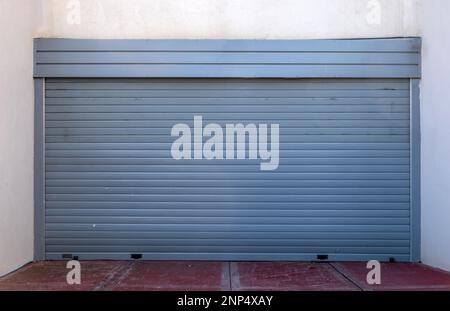 White wall with blue empty roller shutter door background. Metal garage door construction, closed foldable entrance, red entry floor, safety. Space Stock Photo