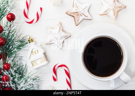 A white mug of black Americano coffee with Spruce branch tree on a white wooden background next to Christmas cookies, a candy cane and New Year's toys Stock Photo