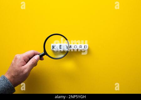 Hand holding a magnifying glass, researching the word keyword on a bright yellow background Stock Photo