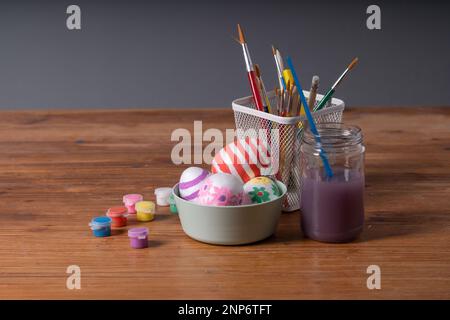 A collection of hand-decorated Easter eggs in a bowl, surrounded by paint jars, brushes, and a pencil holder on a wooden table. Ideal for Easter arts Stock Photo
